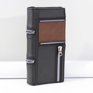 May include: A black faux leather pencil case with a brown panel and silver zippers. The case is designed to resemble a book, with vertical stitching along the spine and a zipper closure on the front.