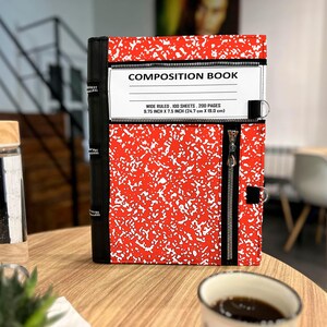 May include: A red and white speckled composition book cover featuring a black spine, a horizontal zipper pocket, and a white label printed with COMPOSITION BOOK, WIDE RULED, 100 SHEETS, 200 PAGES, 9.75 INCH X 7.5 INCH (24.7 cm X 19.0 cm).