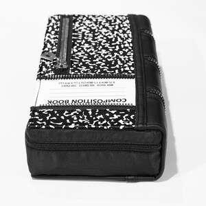 May include: Black composition book-style clutch bag with a zipper closure and a white and black speckled pattern. The bag has a label that reads "COMPOSITION BOOK" and "WIDE RULE 100 SHEETS 20D PAGES 8.75 INCH X 7.5 INCH (22.22cm X 19.05cm)"