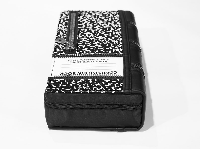 May include: Black composition book-style clutch bag with a zipper closure and a white and black speckled pattern. The bag has a label that reads "COMPOSITION BOOK" and "WIDE RULE 100 SHEETS 20D PAGES 8.75 INCH X 7.5 INCH (22.22cm X 19.05cm)"