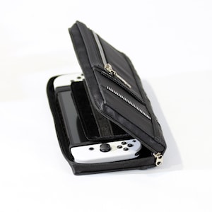 May include: A black zippered case with a white Nintendo Switch console and a black Joy-Con controller inside. The case has a zippered pocket on the front.