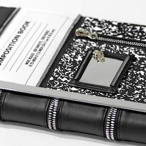 Könnte beinhalten: Ein schwarz-weißes Notizbuch-Cover mit gesprenkeltem Muster, einer Reißverschlusstasche und einem kleinen rechteckigen Spiegel. Der Text lautet COMPOSITION BOOK, WIDE RULED, 100 SHEETS, 200 PAGES, 9.75 INCH X 7.5 INCH (24.7 cm X 19.0 cm).