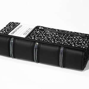 May include: Black zippered book-shaped clutch with a composition notebook pattern and silver zipper pulls. The front of the clutch has a clear plastic window with the text "COMPOSITION BOOK" printed on a white label.