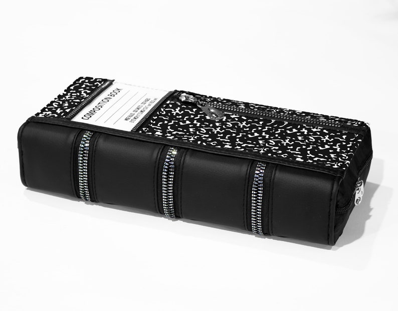 May include: Black zippered book-shaped clutch with a composition notebook pattern and silver zipper pulls. The front of the clutch has a clear plastic window with the text "COMPOSITION BOOK" printed on a white label.