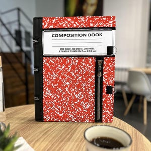 Könnte beinhalten: Ein rot-weißes Composition Book mit schwarzem Rücken und Reißverschluss. Das Cover zeigt den Text "COMPOSITION BOOK" und Details: "WIDE RULED. 100 SHEETS. 200 PAGES 24,7 cm X 19,0 cm".