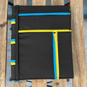 May include: A black rectangular portfolio case featuring multiple exterior pockets secured with bright blue and yellow zippers. The front panel includes a horizontal yellow zipper and a vertical yellow zipper, while the spine has blue and yellow zipper accents.