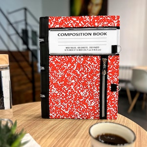 May include: A red and white speckled composition book cover with a black spine and black zipper accents. The white label reads COMPOSITION BOOK, WIDE RULED, 100 SHEETS, 200 PAGES, 9.75 INCH X 7.5 INCH. It features a vertical zipper and metal rings.