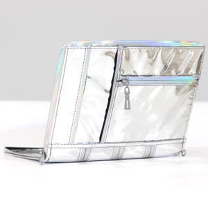 May include: A rectangular, metallic silver pouch with a high-gloss, iridescent finish. The pouch features a horizontal zippered pocket on the front, vertical stitched paneling, and a silver zipper pull tab. The material has a reflective surface.