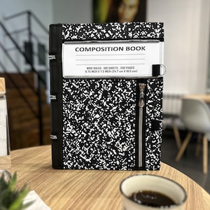 May include: A black and white composition book with a zipper closure. The cover has a speckled pattern and the text "COMPOSITION BOOK" is visible. The book measures 9.75 inches by 7.5 inches.