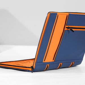 Könnte beinhalten: Eine blaue Laptoptasche mit einer orangefarbenen Reißverschlusstasche und schwarzen Reißverschlüssen. Die Tasche ist geschlossen und der Reißverschluss ist nach oben gezogen.