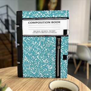 Może przedstawiać: Notatnik z turkusowo-białym nakrapianym wzorem. Okładka posiada zamek błyskawiczny i napis "COMPOSITION BOOK". Książka ma wymiary 24,7 cm x 19,0 cm i ma 200 stron.