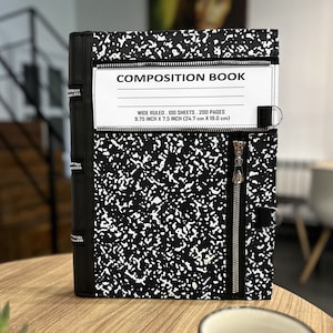 May include: A black and white composition book with a zipper closure. The cover has a speckled pattern. The text "COMPOSITION BOOK" is visible. The book measures 24.7 cm by 19.0 cm.