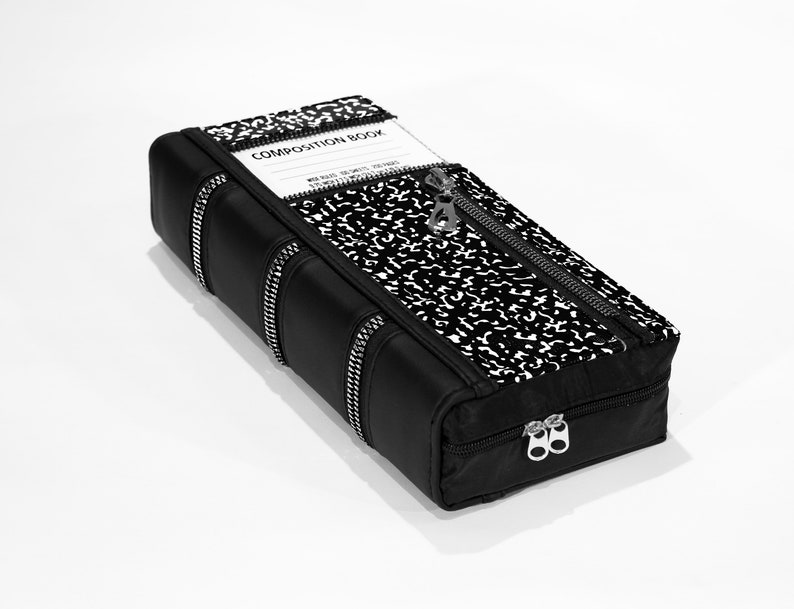 May include: A black zippered pouch shaped like a book with a composition notebook pattern on the front. The pouch has the words "COMPOSITION BOOK" printed on the front.