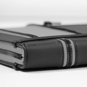 May include: A rectangular black and gray zippered case featuring a smooth, structured exterior. The design includes a prominent black zipper track running along the side and top edges, with gray panels integrated into the black frame construction.
