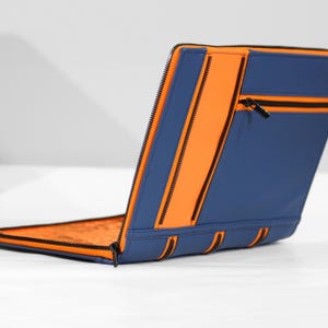 May include: A rectangular portfolio case featuring a vibrant orange and deep blue color scheme. The exterior includes a horizontal zippered pocket with a black pull tab, while the interior lining displays a detailed orange pattern.