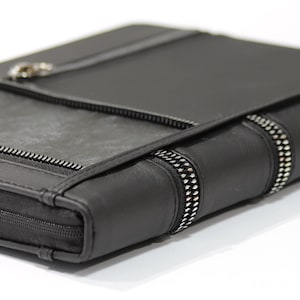 Könnte beinhalten: Eine schwarze Ledermappe mit Reißverschluss und dekorativen Reißverschlussakzenten. Die Mappe hat eine strukturierte Oberfläche und einen silberfarbenen Verschluss. Das Design ist schlicht und modern.