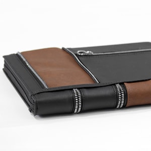 May include: A rectangular portfolio case featuring alternating panels of smooth black and brown material. The design includes multiple silver-toned metal zippers that run horizontally across the front and along the side edge of the case.