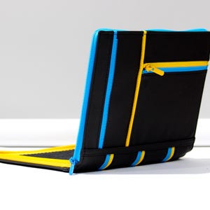 May include: A black rectangular portfolio case featuring multiple bright blue and yellow zippers arranged vertically and horizontally. The case includes a zippered pocket on the exterior and a yellow interior lining visible at the base.