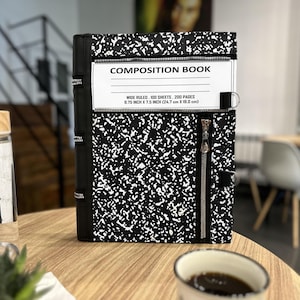 May include: A black and white speckled composition book cover featuring a white label with the text COMPOSITION BOOK, WIDE RULED, 100 SHEETS, 200 PAGES, 24.7 cm x 19.0 cm. The cover includes a vertical zip and a black spine with zip details.