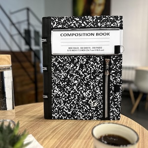 May include: A black and white speckled composition book cover featuring a horizontal zipper pocket across the top and a vertical zipper pocket on the right side. The white label reads COMPOSITION BOOK, WIDE RULED, 100 SHEETS, 200 PAGES, 9.75 INCH X 7.5 INCH.