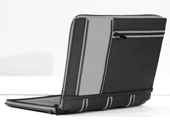 May include: A rectangular portfolio case featuring a two-tone design of black and light gray panels. The exterior includes multiple vertical and horizontal zipper closures with black pulls, providing a structured look with clean, straight lines.