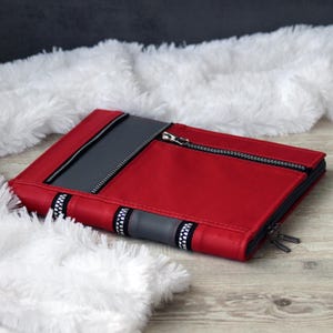 May include: A rectangular red and gray zippered portfolio case featuring a prominent diagonal zipper across the front panel, a secondary horizontal zipper along the edge, and black and white patterned trim accents along the lower section.
