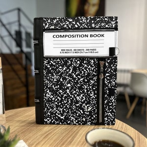Puede incluir: Un cuaderno de composición negro con cierre de cremallera y una etiqueta blanca que dice "COMPOSITION BOOK, WIDE RULED, 100 SHEETS, 200 PAGES, 9.75 INCH X 7.5 INCH (24.7 cm X 19.0 cm)"