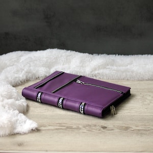 Könnte beinhalten: Eine lila Clutch im Buchstil aus Leder mit silbernen Reißverschlüssen und dekorativen Bändern. Die Clutch ist geschlossen, mit einem Reißverschluss über der Oberseite und einem kleineren Reißverschluss in der unteren rechten Ecke. Der Buchrücken hat schwarze Bänder mit weißen und silbernen Verzierungen.