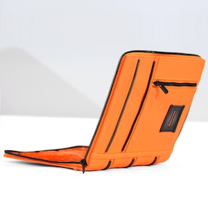 May include: Bright orange zippered case with black trim and a small pocket. The case has a rectangular label that reads "Kelvin Tran". The case is open, revealing the interior.