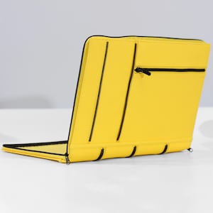 Könnte beinhalten: Eine leuchtend gelbe Laptoptasche mit schwarzen Reißverschlüssen und Akzenten. Die Tasche ist geöffnet und zeigt das Innere. Das Design umfasst mehrere Reißverschlussfächer und eine elegante, moderne Ästhetik. Das Produkt besteht aus einem glatten, strapazierfähigen Material.