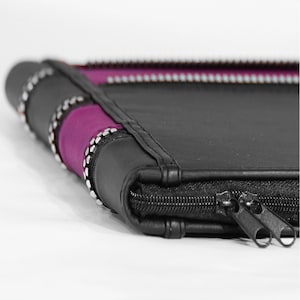 Könnte beinhalten: Ein schwarzes Leder-Zipper-Portfolio mit einem violetten und silbernen Besatz. Das Portfolio hat ein dekoratives Design mit silbernen Nieten und einem Reißverschluss.