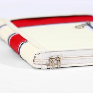 May include: A rectangular off-white case featuring a prominent red horizontal stripe and a secondary red accent band. The case is constructed with a textured off-white fabric border and includes two silver-toned metal zipper pulls on the side.