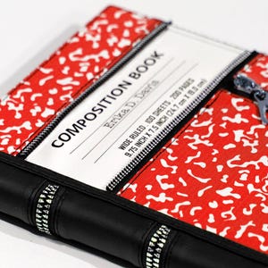 May include: A red and white speckled composition book cover featuring a black spine with zipper accents. The white label reads COMPOSITION BOOK, Erika D. Desha, WIDE RULED, 100 SHEETS, 200 PAGES, 9.75 INCH X 7.5 INCH. A silver zipper pull is visible.