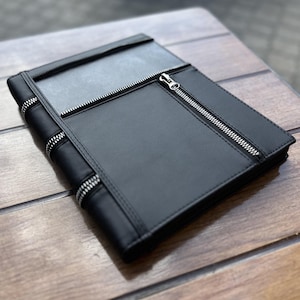 May include: A black rectangular portfolio case featuring a smooth matte finish. The front panel includes a prominent silver-toned metal zipper that runs diagonally across the surface, complemented by additional decorative zipper accents along the side.