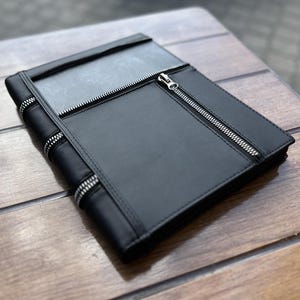 May include: A black rectangular portfolio case featuring a smooth surface with multiple silver-toned metal zippers. The design includes a vertical zipper closure on the front panel and additional zipper detailing along the side edge of the cover.