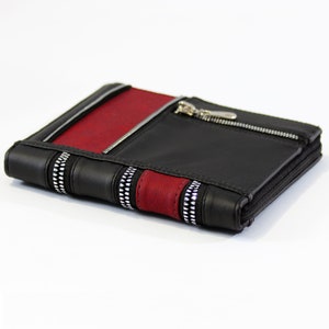 May include: A rectangular black wallet featuring a vertical red stripe and silver-toned zipper accents. The wallet includes a zippered pocket on the front, multiple stitched panels, and a wrap-around zipper closure along the outer edge.