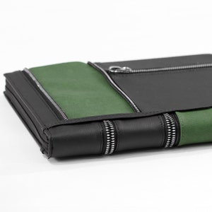 May include: A rectangular black and green wallet featuring multiple silver-toned metal zippers. The exterior is constructed from smooth black panels contrasted with textured green sections, with visible zipper teeth along the edges and front.