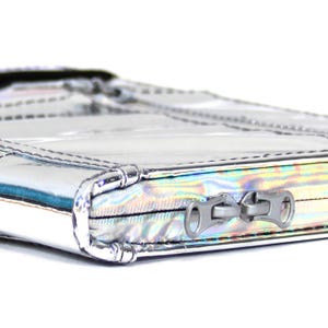 May include: A rectangular silver metallic pouch with a holographic finish and a quilted texture. The pouch features two silver-toned metal zipper pulls attached to a zipper closure that runs along the side of the item, showcasing stitched seams.