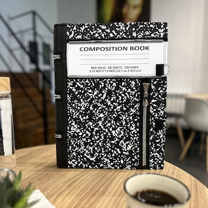 May include: Black composition book with a zipper closure and a white label that reads "COMPOSITION BOOK, WIDE RULED, 100 SHEETS, 200 PAGES, 9.75 INCH X 7.5 INCH (24.7 cm X 19.0 cm)"