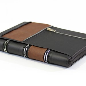 May include: A rectangular portfolio case featuring a combination of black and brown panels. The exterior includes multiple silver-toned metal zippers, including a horizontal zippered pocket on the front and vertical zipper accents along the spine.