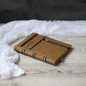 Puede incluir: Una carpeta o funda para cuaderno rectangular de color tostado con una superficie exterior lisa. El diseño incluye detalles de cremallera negra en la parte superior y lateral, con un adorno de cinta estampada en blanco y negro en el lomo.