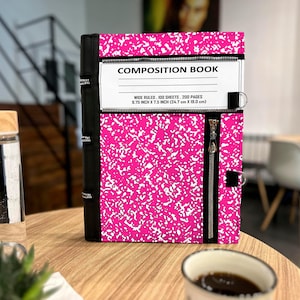May include: A bright pink and white speckled composition book cover featuring a black spine, a horizontal zipper across the top, and a vertical zipper on the right side. The label reads COMPOSITION BOOK, WIDE RULED, 100 SHEETS, 200 PAGES, 9.75 INCH X 7.5 INCH.