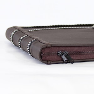 Puede incluir: Un estuche rectangular de color marrón oscuro con superficie lisa y cierre de cremallera. El lateral presenta tres bandas decorativas con un patrón en blanco y negro. Dos tiradores de cremallera de metal negro están unidos al cierre.