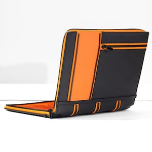 May include: A black and orange laptop case with a zipped pocket on the back. The case has a zipped closure and a soft, padded interior.