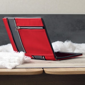 May include: A rectangular portfolio case featuring a vibrant red and dark gray color scheme. The exterior includes a prominent horizontal zipper pocket with silver-toned teeth and a matching zipper pull, accented by vertical gray panels.
