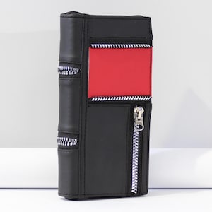May include: Black wallet with a red panel and silver zippers. The wallet is designed to resemble a book, with vertical lines and a zipper closure. The zippers have a silver finish, and the wallet is made of a smooth material.