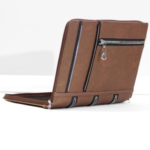 May include: A brown rectangular portfolio case featuring multiple silver-toned metal zippers. The exterior has a smooth finish with one horizontal zipper pocket at the top and two vertical zippers running down the front panel, secured by a side zipper.