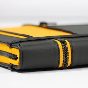 Könnte beinhalten: Nahaufnahme einer schwarzen und gelben Reißverschlusstasche. Die Tasche hat eine schwarze Außenseite mit gelben Akzenten und einen auffälligen gelben Reißverschluss. Die Zähne des Reißverschlusses sind schwarz. Die Tasche scheint aus Leder oder einem ähnlichen Material zu bestehen.