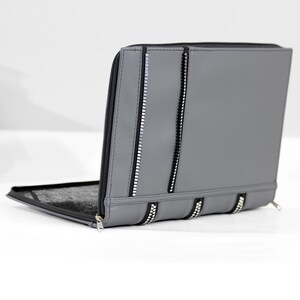 May include: A gray zippered portfolio case featuring a smooth exterior surface with two vertical black and silver decorative zipper accents on the front panel and a black interior lining visible on the open side of the rectangular document holder.