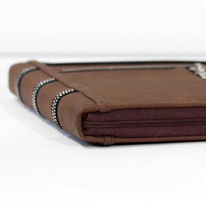 May include: A brown leather wallet with a zipper closure. The wallet has a decorative silver and black trim on the spine and a matching zipper. The wallet is rectangular and appears to be made of a durable material.